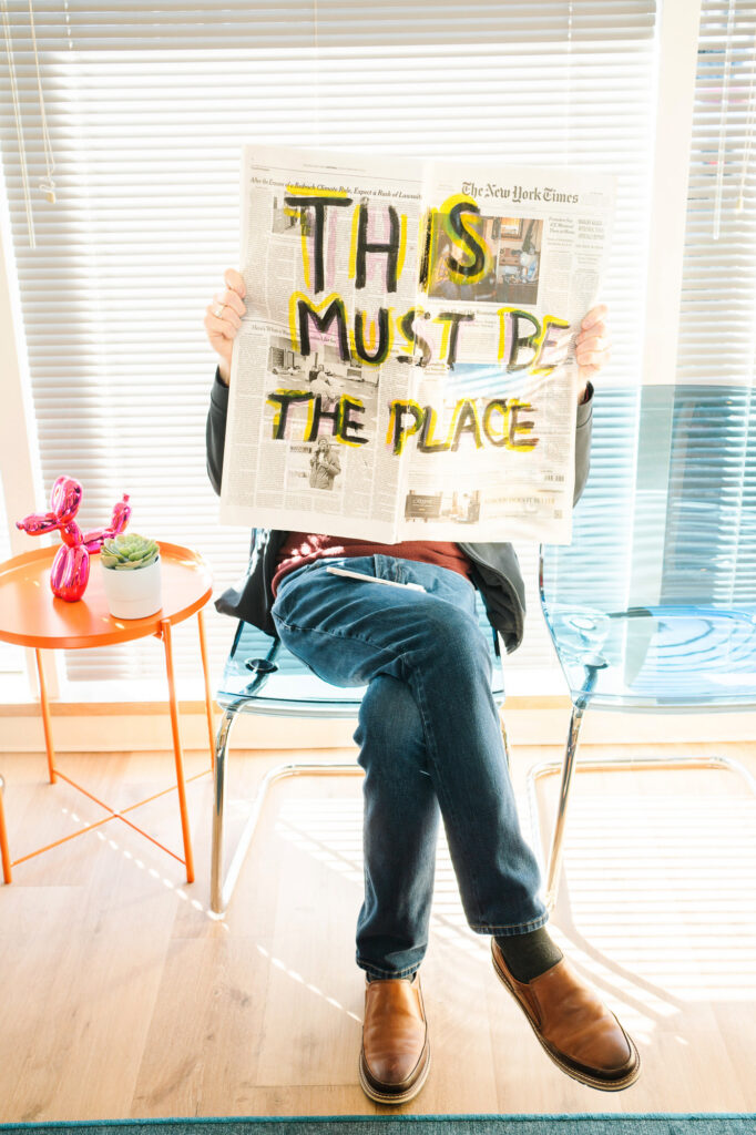 Brand photo of a man sitting in a waiting room holding up a newspaper that that has the words This Must Be The Place written in marker over it. 