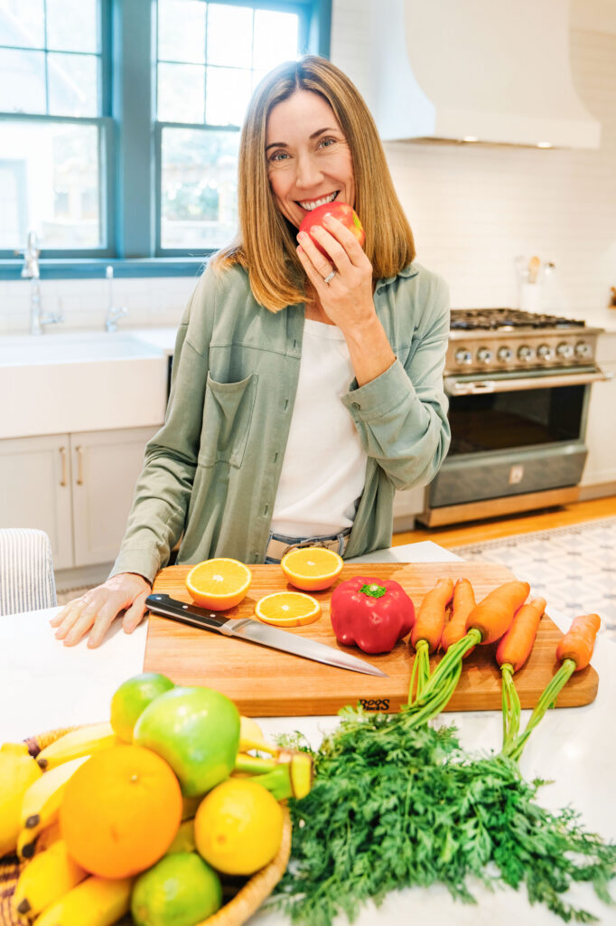 Nutritionist standing in a kitchen in front of a bunch of fruits and vegetable holding an apple to her mouth as if she's going to bite into it. 