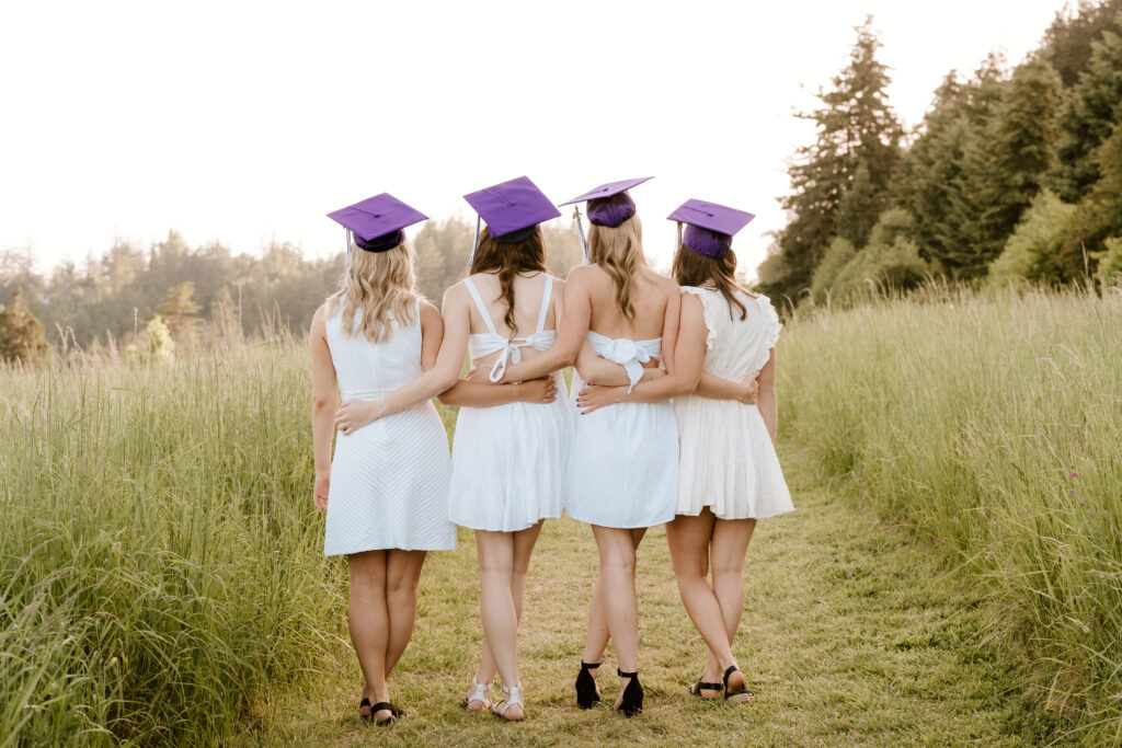 Four high school girls with their backs turned wearing white dresses and their graduation caps during their professional graduation photos.